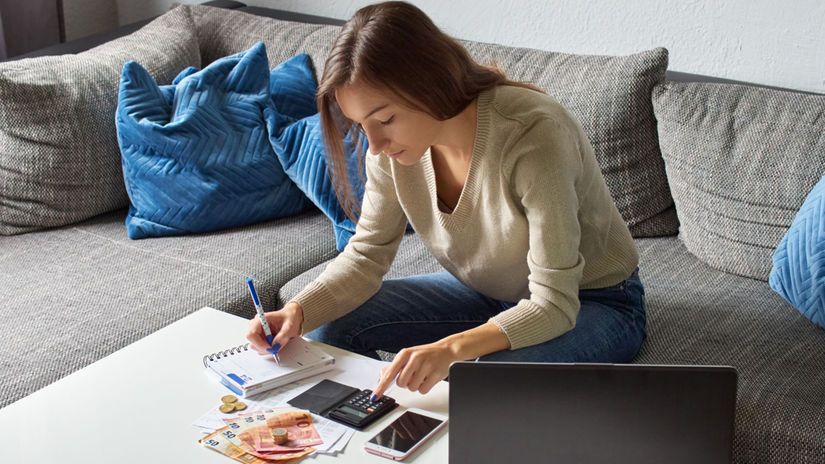 Mulher sentada no sofá fazendo contas com calculadora, anotações e dinheiro sobre a mesa, representando o planejamento financeiro e práticas de como economizar mensalmente com disciplina e organização.