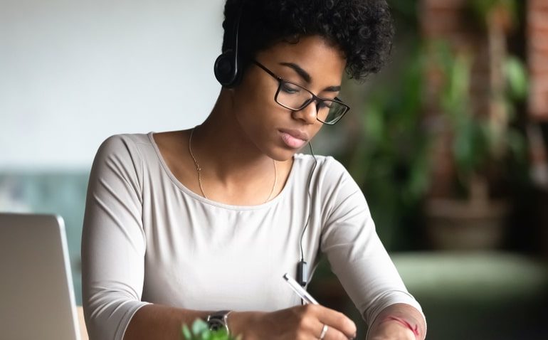 Mulher usando fones de ouvido e óculos enquanto faz anotações em um caderno ao lado de um notebook, representando o planejamento financeiro e o estudo sobre previdência privada.
