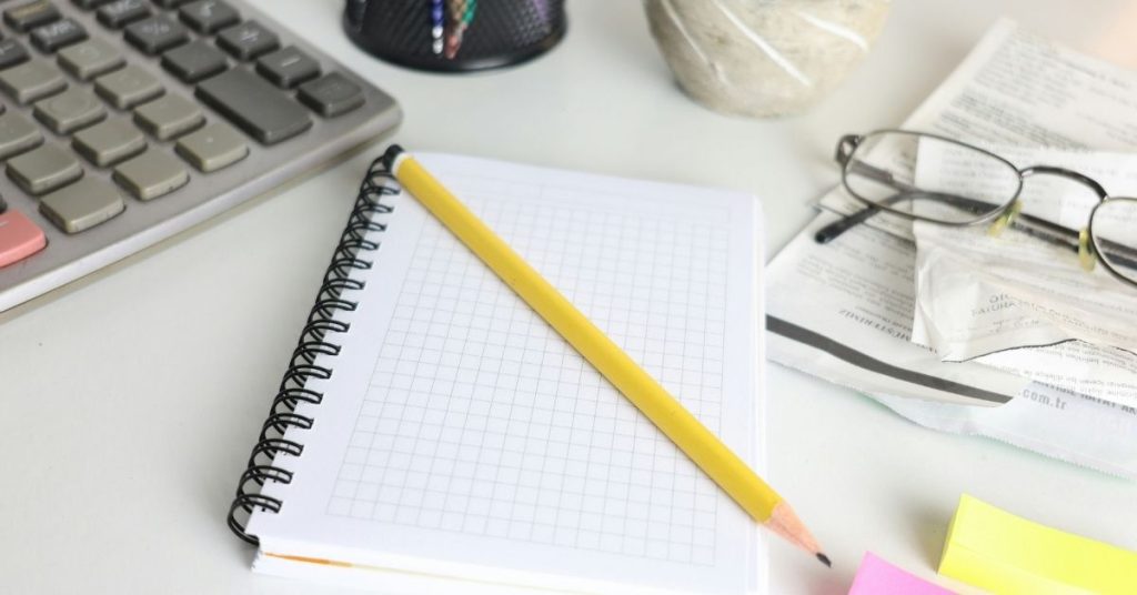 Caderno aberto com lápis amarelo sobre a mesa ao lado de calculadora e contas, representando organização do planejamento financeiro