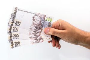 Mão segurando várias cédulas de duzentos reais com destaque para o valor impresso e o rosto da efígie da República, representando o pagamento do décimo terceiro salário.