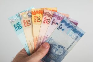 Mão segurando cédulas de real brasileiro em diferentes valores, incluindo notas de 2, 5, 10, 20, 50 e 100 reais, representando dinheiro físico e o contexto financeiro do décimo terceiro salário.