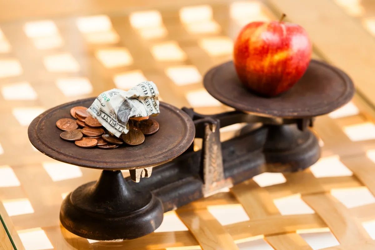 Balança antiga com moedas e dinheiro de um lado e uma maçã do outro, simbolizando como a inflação impacta o poder de compra dos alimentos.