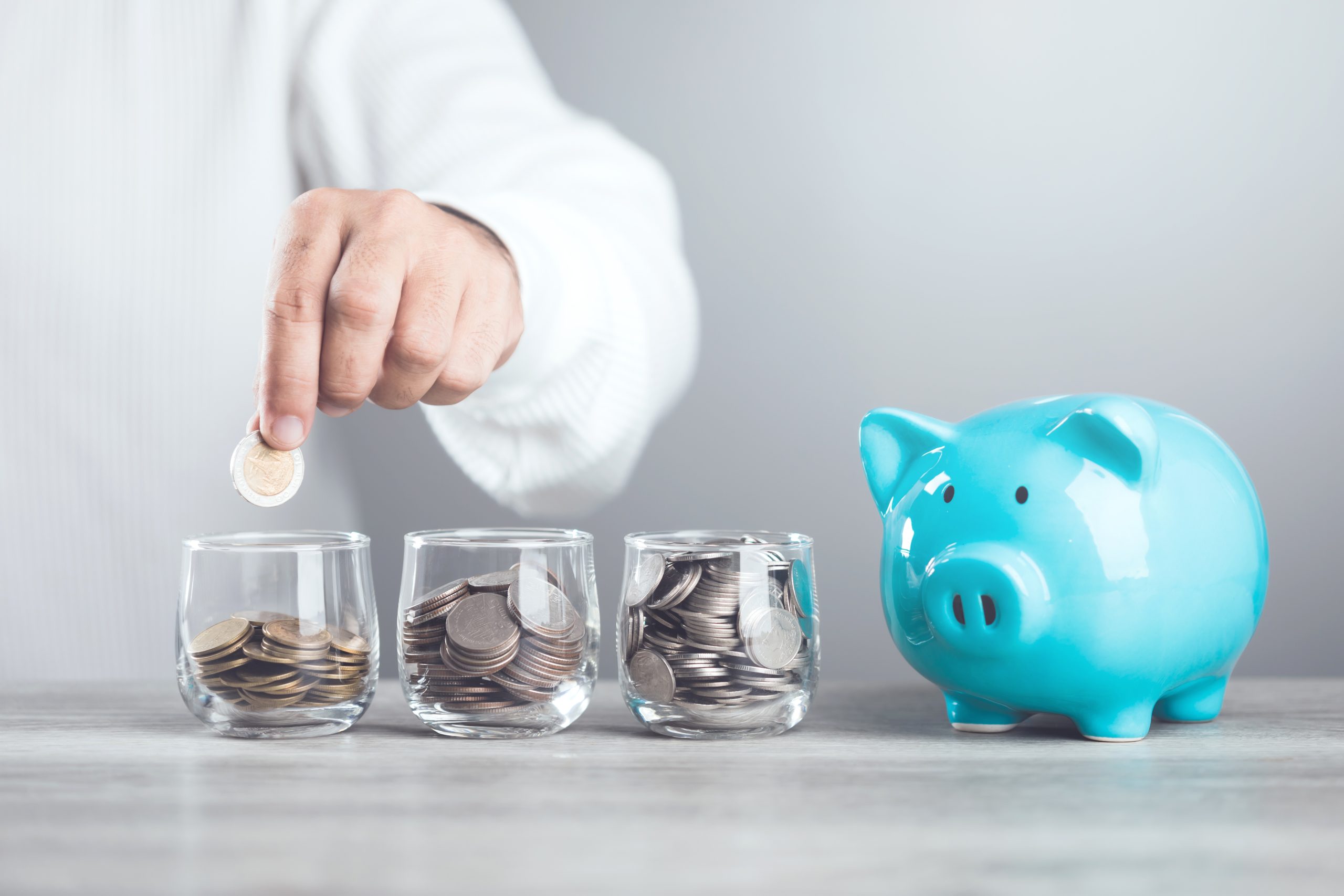Mão colocando moedas em copos de vidro ao lado de um cofrinho azul, simbolizando organização do dinheiro e dicas para melhorar o controle financeiro no dia a dia.