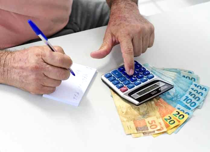 Pessoa calculando valores com calculadora e caderno, ao lado de notas de real, representando empréstimo pessoal para negativado e planejamento financeiro.