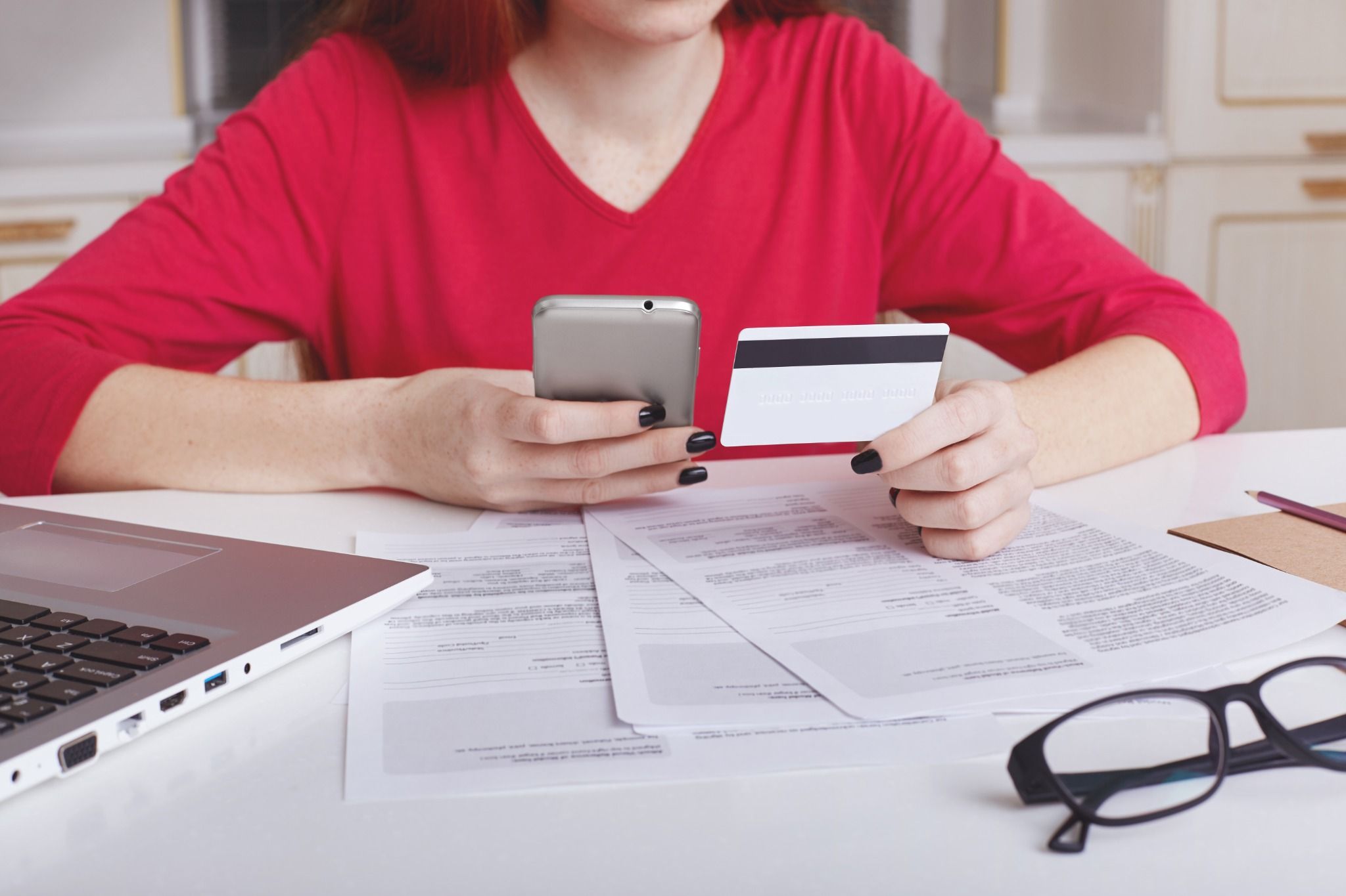 Mulher analisando faturas e segurando cartão de crédito e celular sobre a mesa, refletindo se pagamento mínimo da fatura vale a pena diante das contas acumuladas.