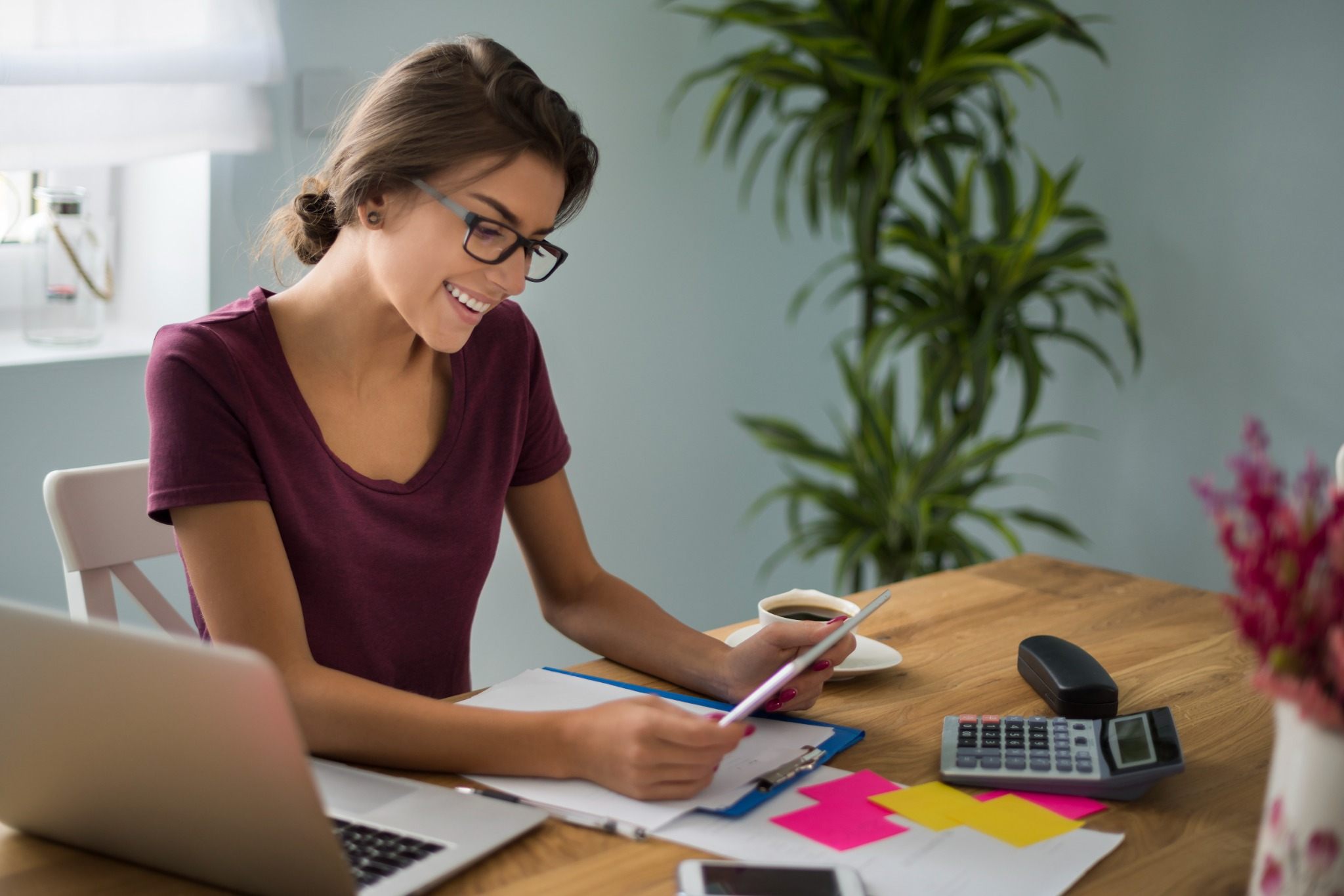 Mulher sorrindo enquanto organiza contas e calcula despesas em casa, representando planejamento financeiro pessoal simples com laptop, calculadora e anotações sobre a mesa.